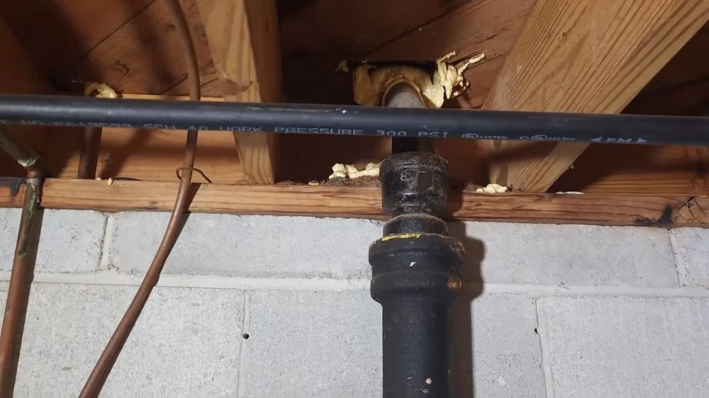 Black cast iron drain joined to black plastic pipe under wooden joists above a block wall