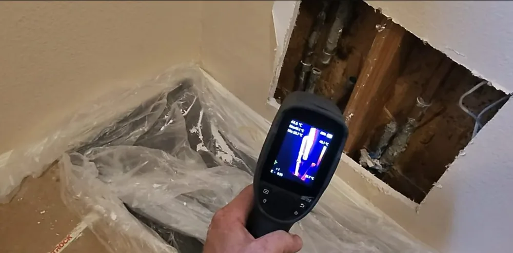 Hand holding a thermal imaging camera toward an opened section of drywall, with plastic sheeting on the floor below