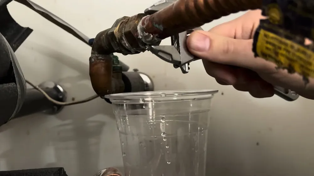 Hand using a wrench on the copper inlet of a water heater, with water dripping into a clear plastic cup