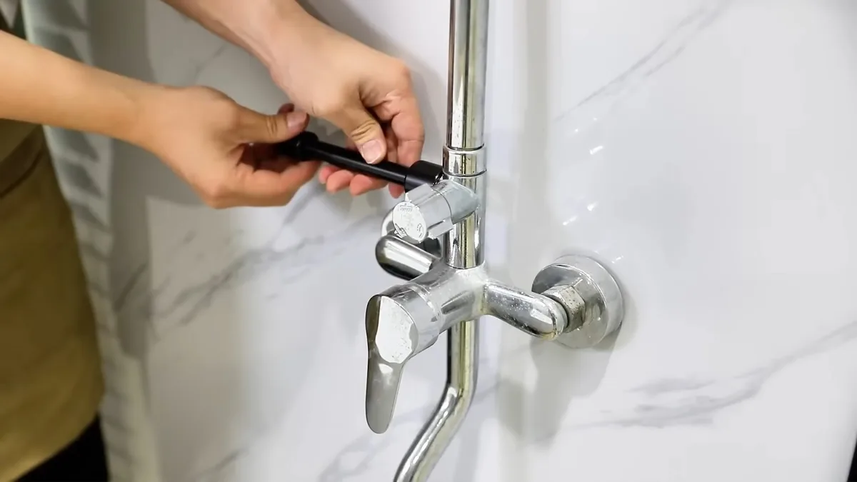 Hands tightening a small screw on a chrome wall-mounted tub and shower faucet against a white marble wall