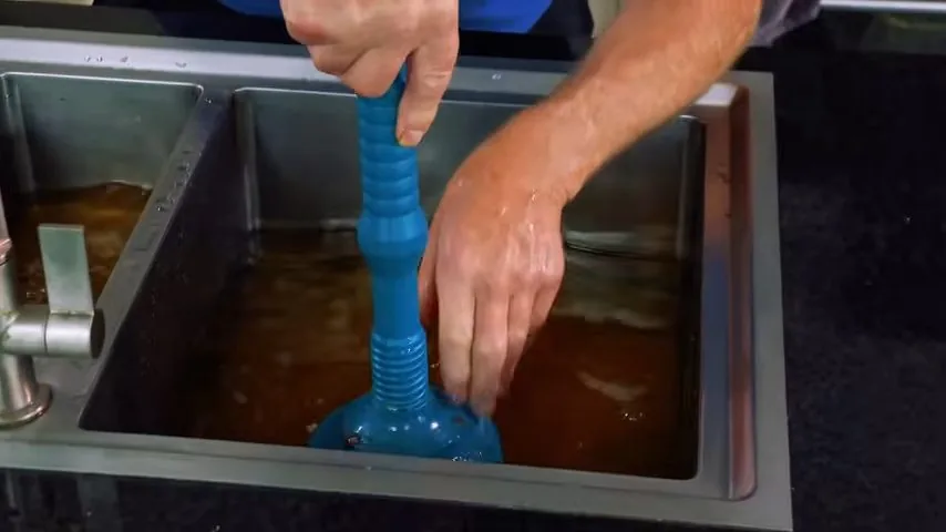 Hands working a blue plunger in a stainless steel sink filled with brown, standing water
