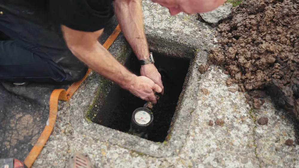Man crouching at a concrete meter box in a driveway, reading the water meter inside it with an orange strap hanging nearby