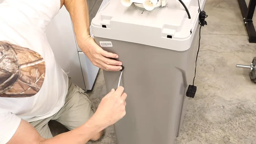 Man in a camouflage cap lifting a gray water softener cabinet onto its stand