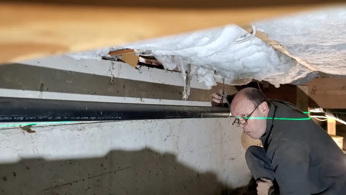 Man in glasses kneeling at a basement wall beside a black pipe along the ceiling, a green laser line crossing the frame