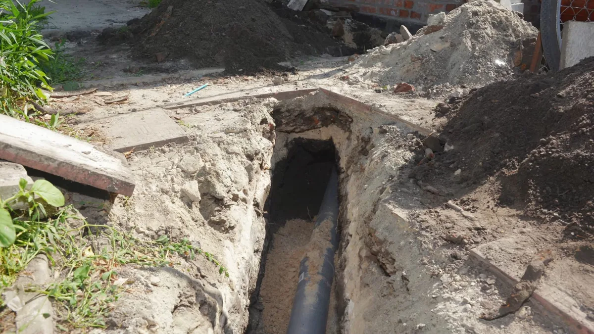 Open trench running toward a house, with excavated soil piled on both sides and a dark pipe at the bottom
