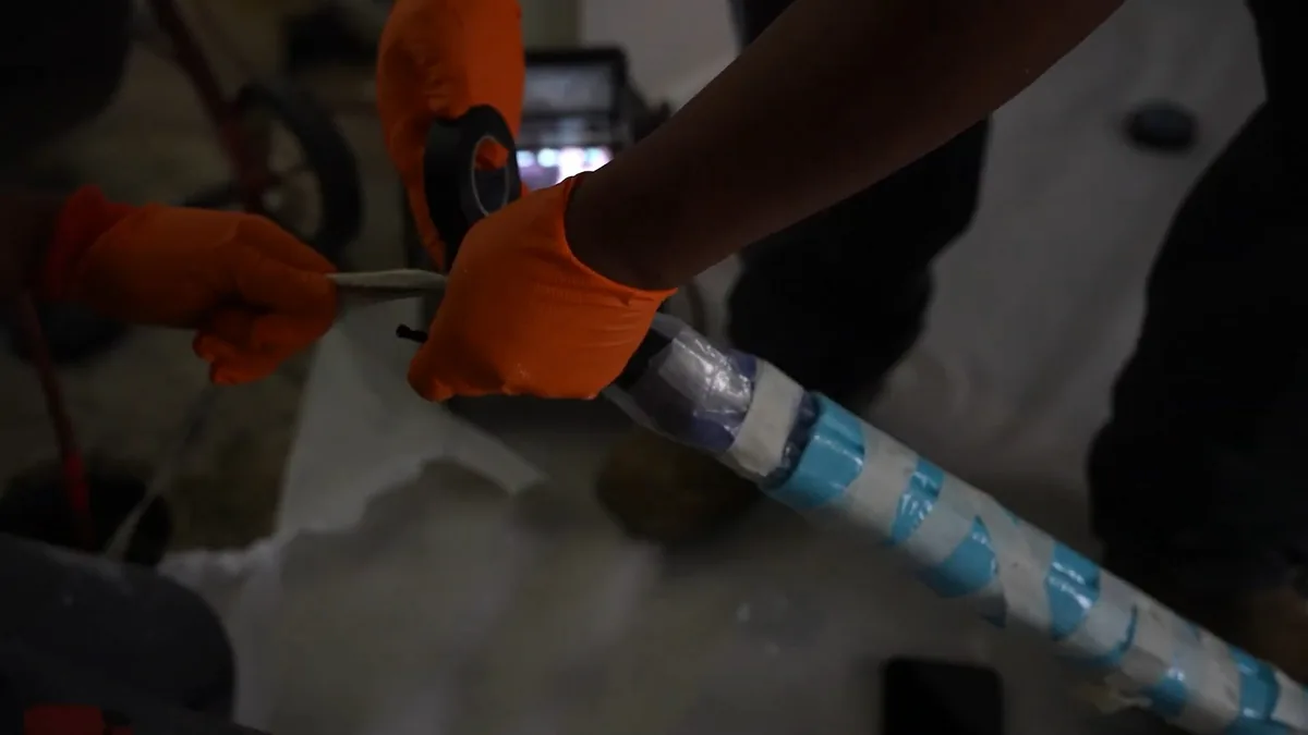 Orange-gloved hands pulling a blue-wrapped liner tube along the floor beside a worker