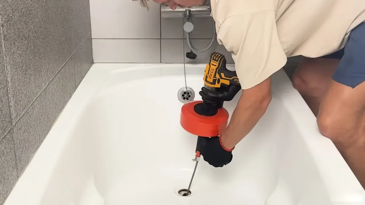 Person using a cordless drill to spin a red drain auger down a bathtub drain