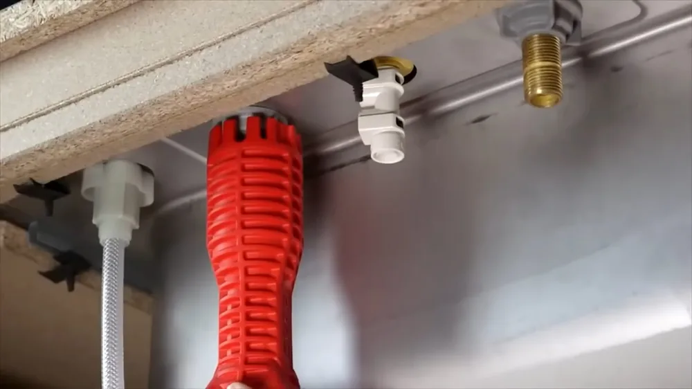 Red multi-tool wrench resting under a sink beside the supply lines and drain fittings