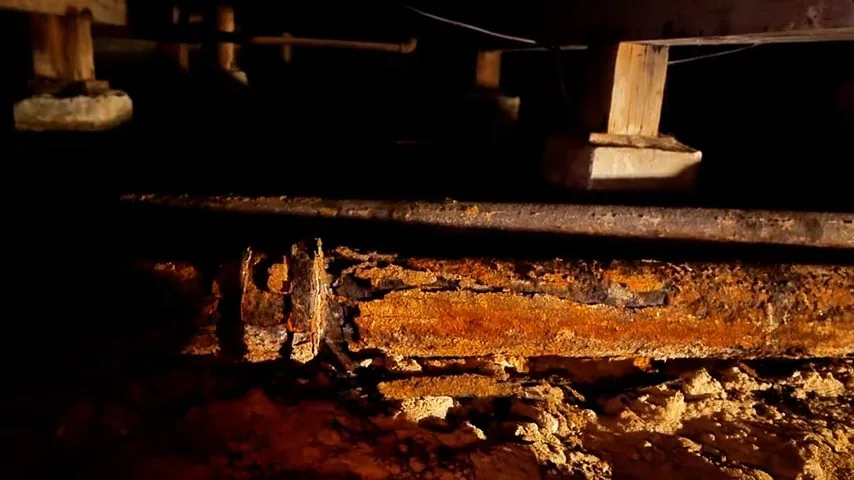 Rusted cast iron sewer pipe beneath wooden floor joists in a dark crawl space