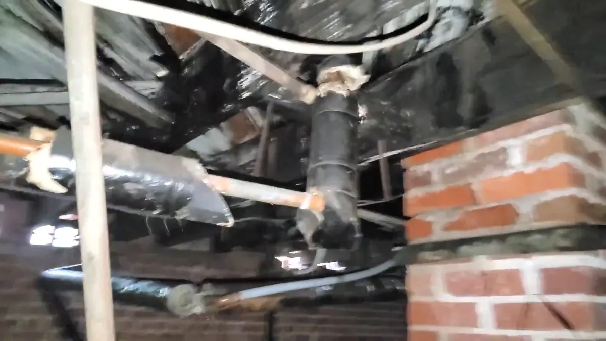 Rust-streaked pipe joint in a crawl space beside a brick foundation wall, with moisture around the fitting