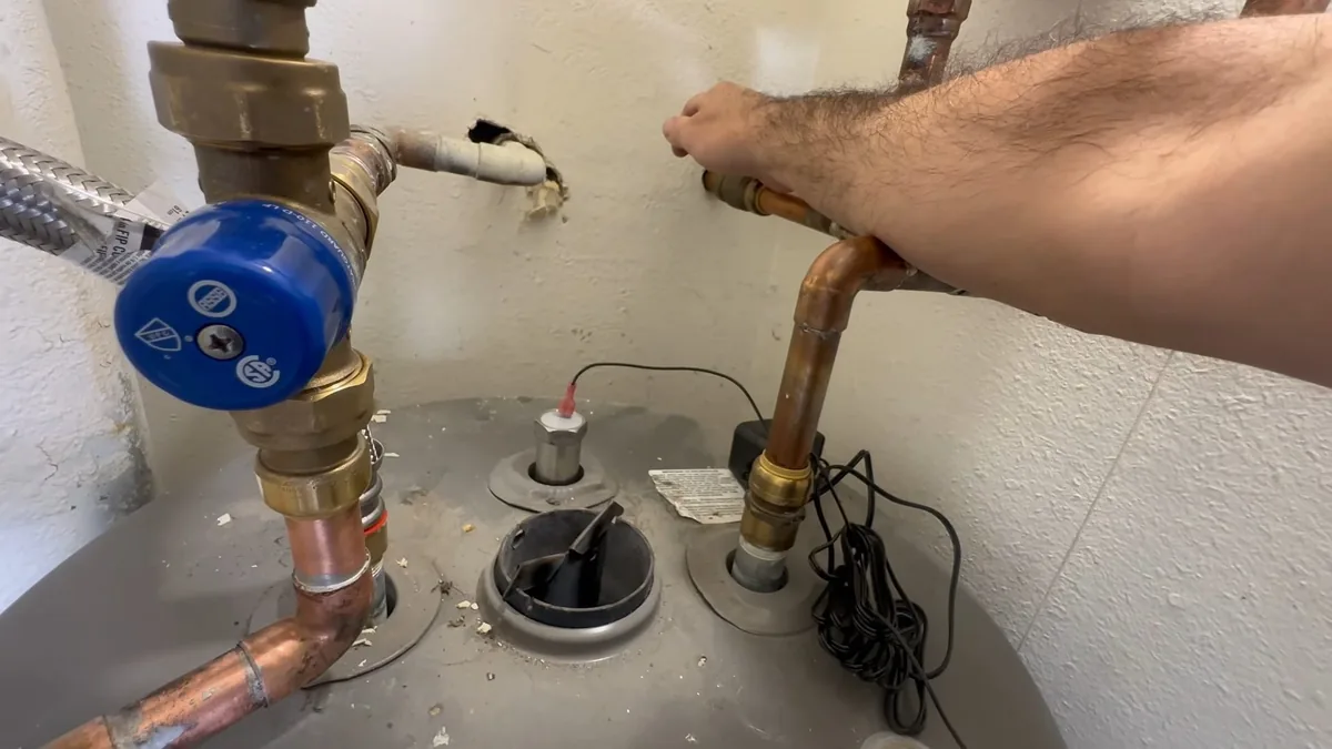 Technician's hand resting on copper supply lines above a tank water heater, beside a blue shutoff valve