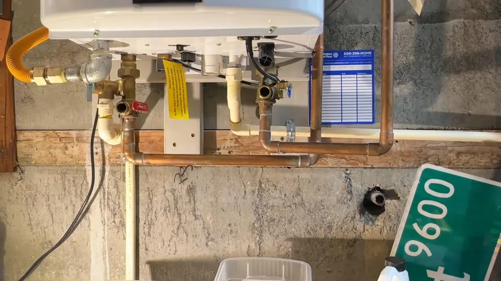 Tankless water heater mounted above copper pipes and a yellow gas line, with a green street sign leaning below