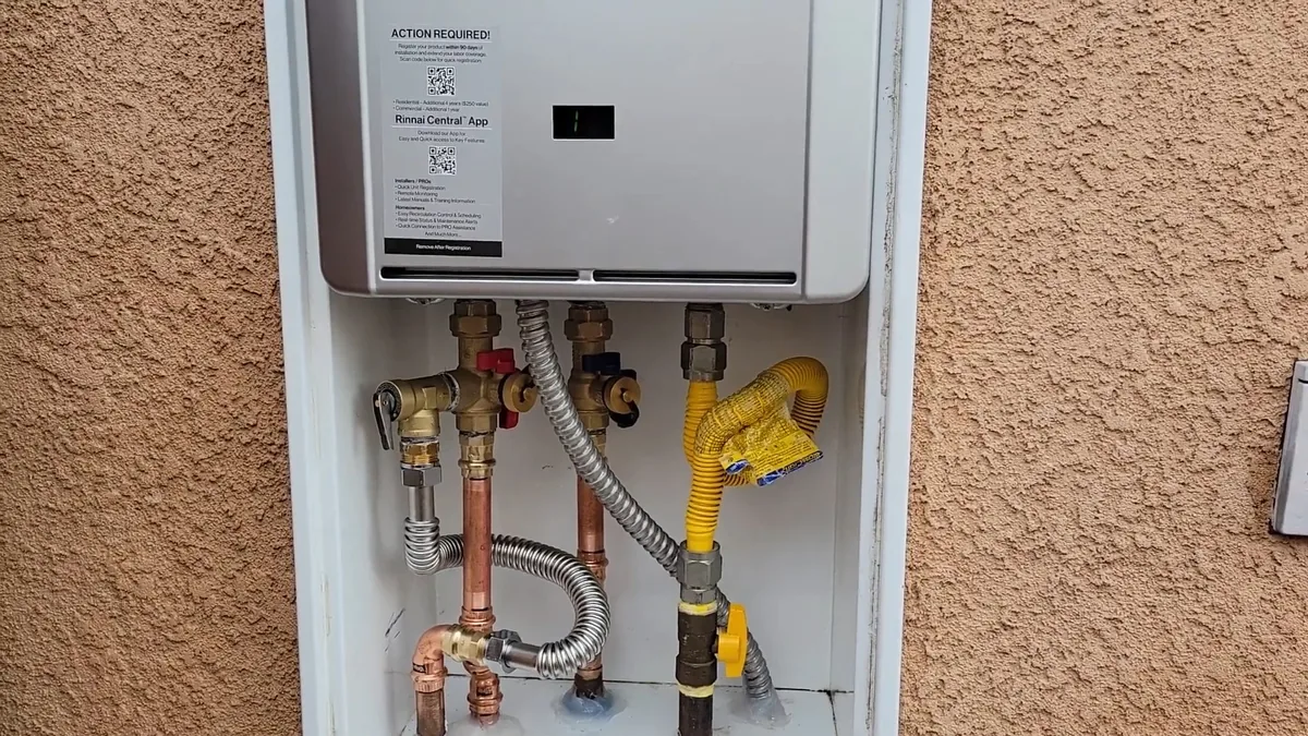 Tankless water heater mounted in an open wall cabinet on a stucco exterior, with gas and water lines below