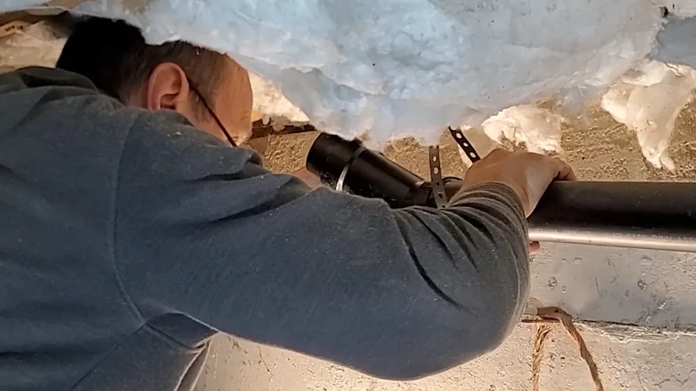 Technician reaching up to a black drain line beneath an insulated floor in a crawl space
