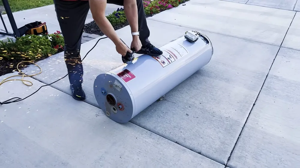 Two people moving a white tank water heater lying on its side across a concrete patio