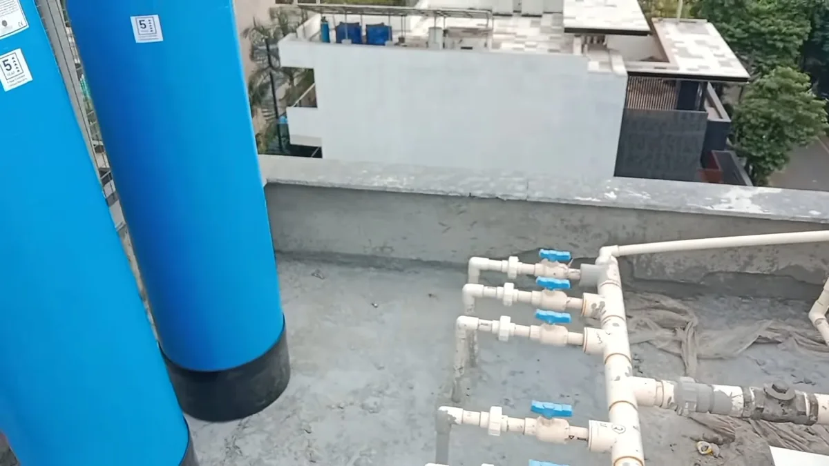 Two tall blue water softener tanks and a white PVC manifold with blue valves on a flat roof