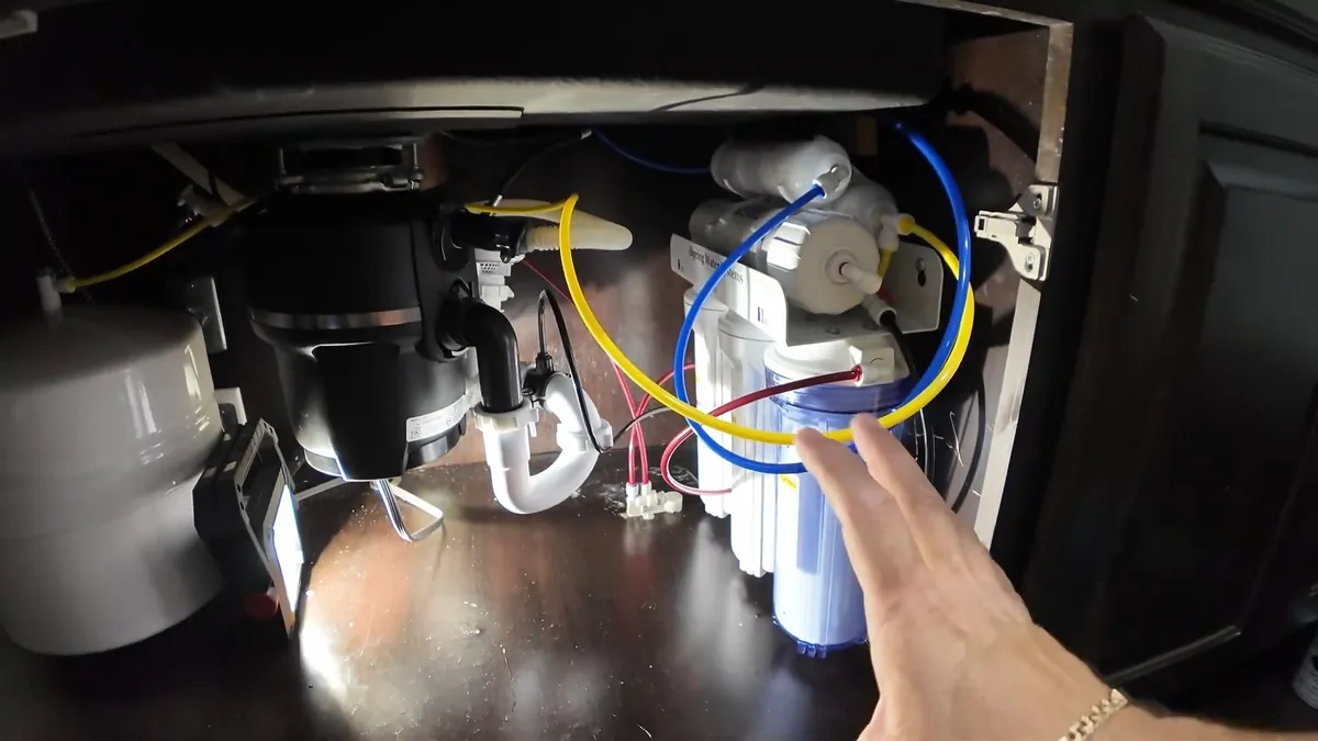 Under-sink cabinet with a garbage disposal mounted at the drain and a multi-stage water filter beside it, a hand pointing at the fittings
