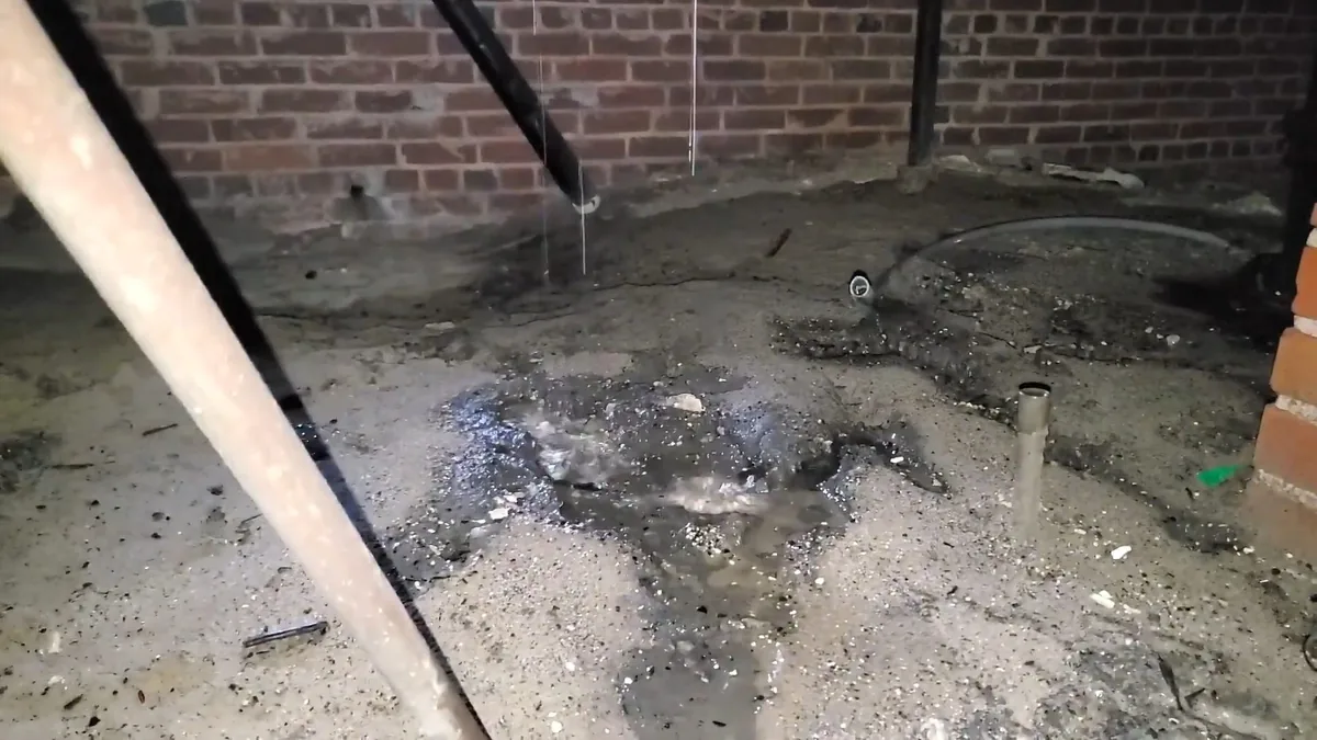 Wet, stained concrete floor beside a brick wall with a pipe stub rising from the ground