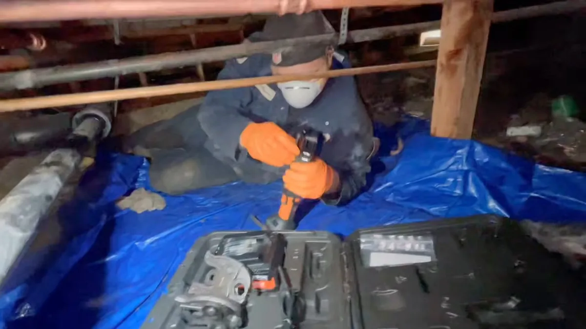 Worker in a dust mask and orange gloves repairing a leaking pipe in a crawl space, kneeling on a blue tarp beside a toolbox