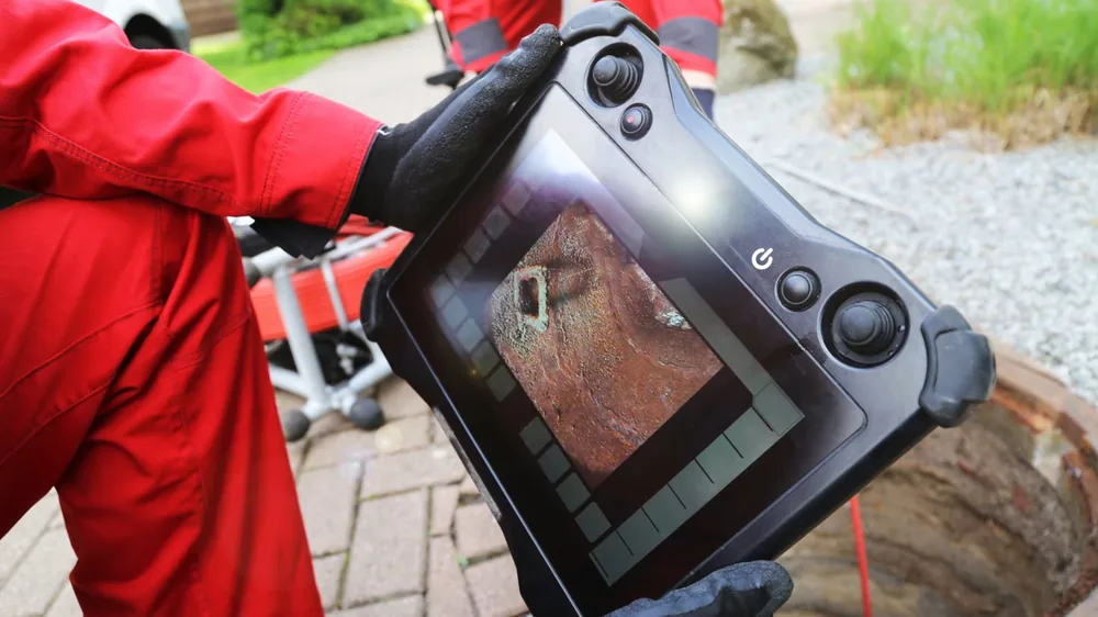 Worker in a red jacket holding a handheld inspection monitor beside an open cleanout, screen showing the inside of a pipe